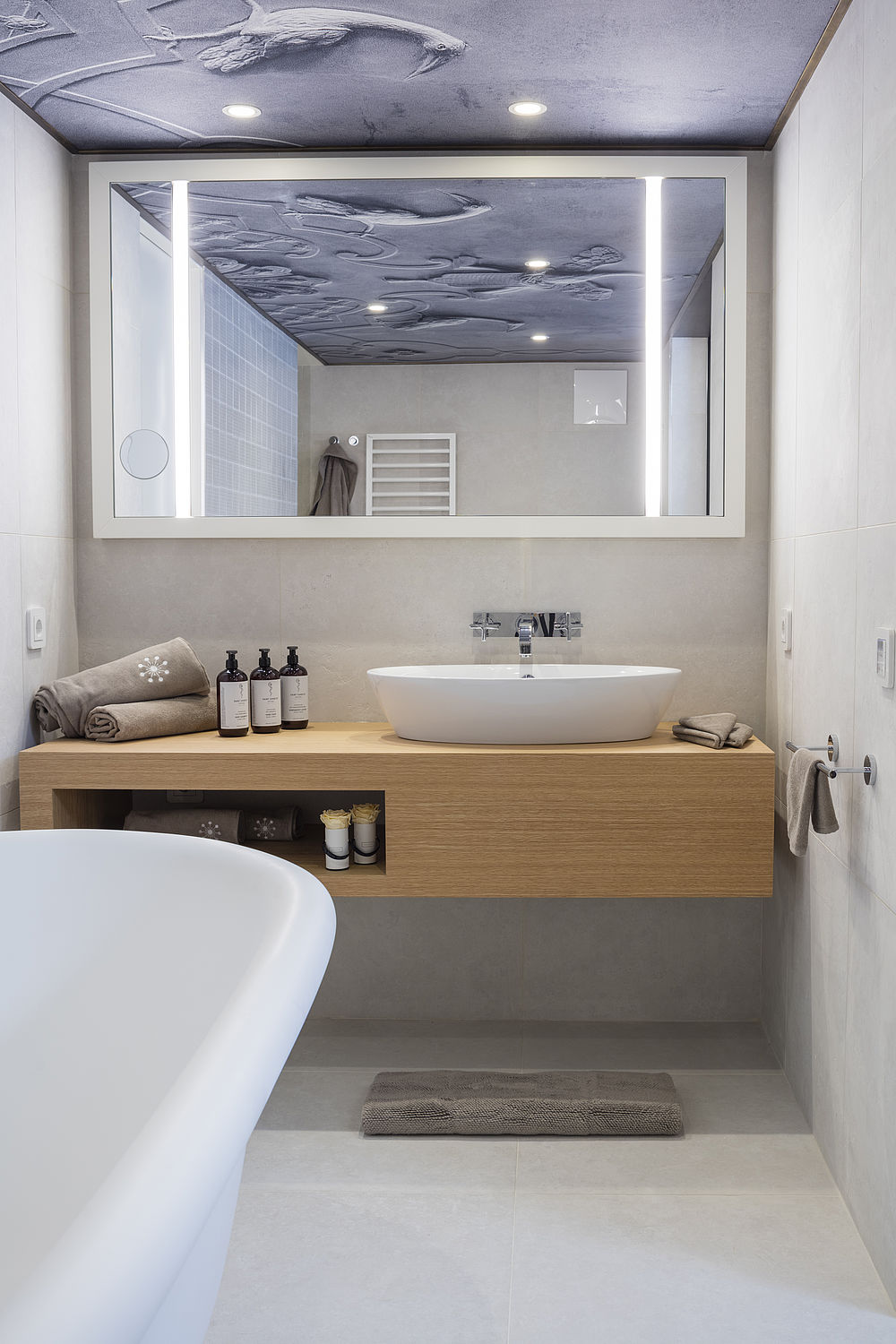 Helles Badezimmer mit großem Spiegel, freistehender Badewanne und bereitgestellten Handtüchern und Pflegeprodukten auf dem Waschbecken