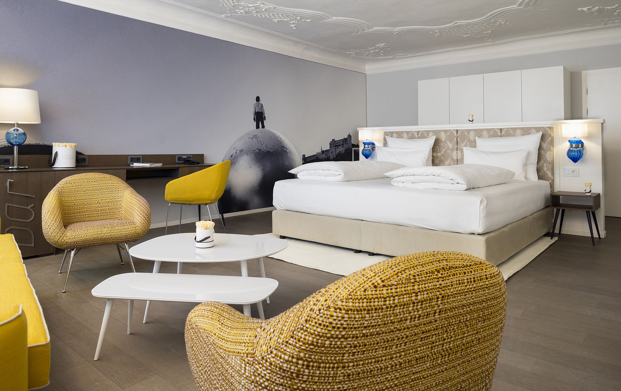 Geräumige Hotel-Suite mit großem Boxspringbett, Sitzecke mit gemütlichen Lounge-Sesseln und Sofa, sowie modernem Design der Wandtapete