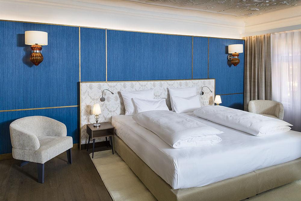 Großes Boxspringbett der Honeymoon Suite vor blauer Rubelli Tapete mit goldenen Verzierungen, zwei kleinen Sesseln und zwei Nachttischen