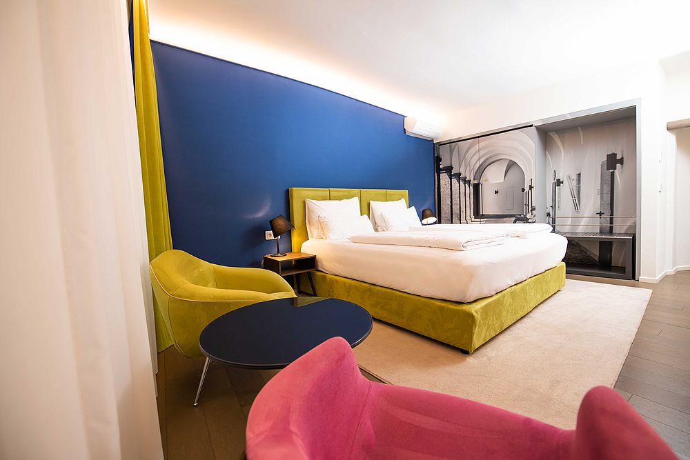 Gelbes Boxspringbrett vor blauer Wand und Sitzecke mit Polstermöbeln in kräftigen Farben in der Junior Suite des Hotel Stein