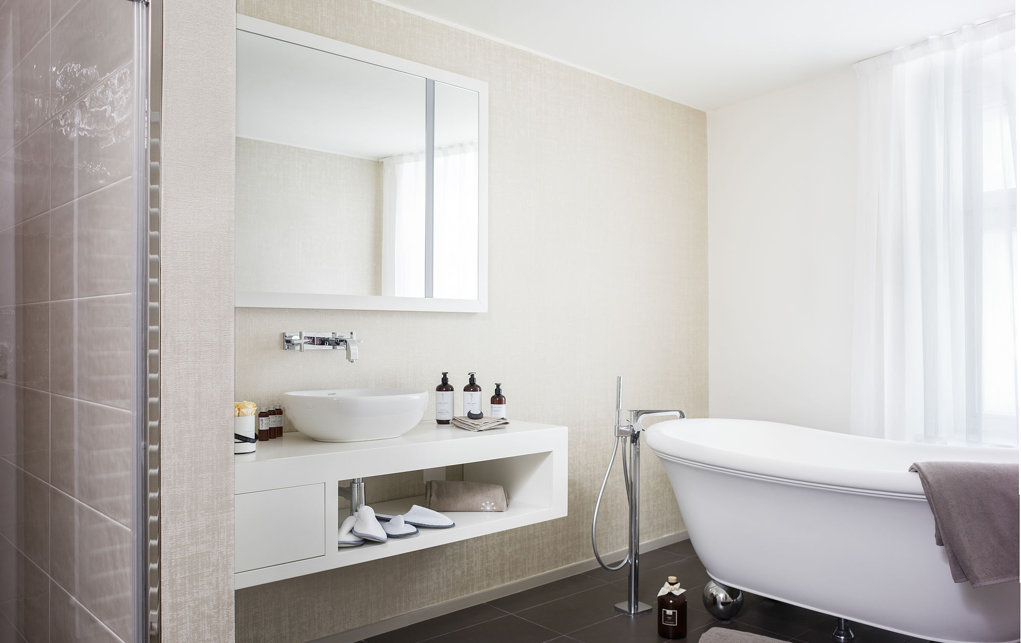 Blick auf Badewanne und Waschbecken einer Hotel Suite mit bereitstehenden Handtüchern, Pflegeprodukten und Hausschuhen