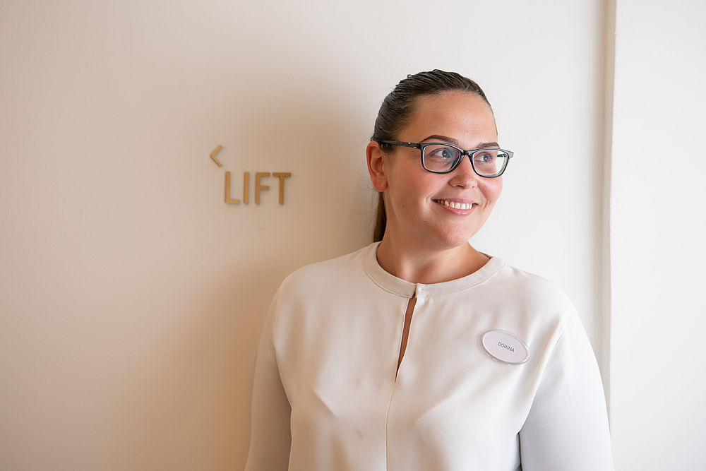 Eine Hotelmitarbeiterin steht lächelnd vor einer Wand mit goldenem Wegweiser zum Aufzug 