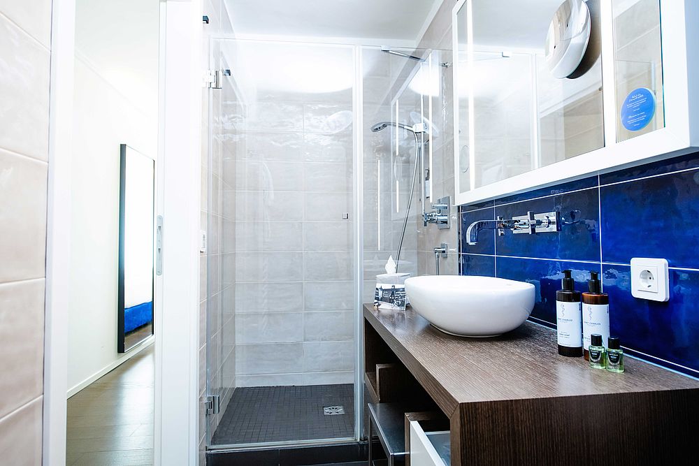Badezimmer im Doppelzimmer des Hotel Stein mit blauen Fließen, Naturkosmetik auf der modernen Waschtischkonsole aus dunklem Holz und einer Glas-Duschkabine