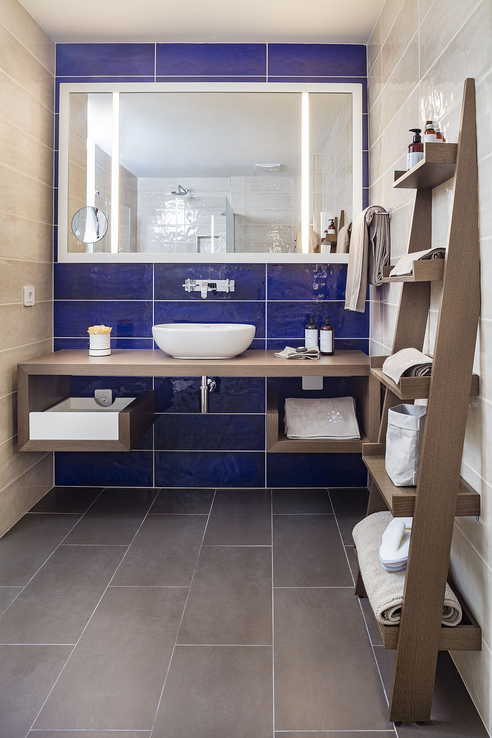 Das Standard-Badezimmer besitzt Möbel aus dunklem Holz, blaue und braune Fliesen, einige Handtücher und Pflegeprodukte
