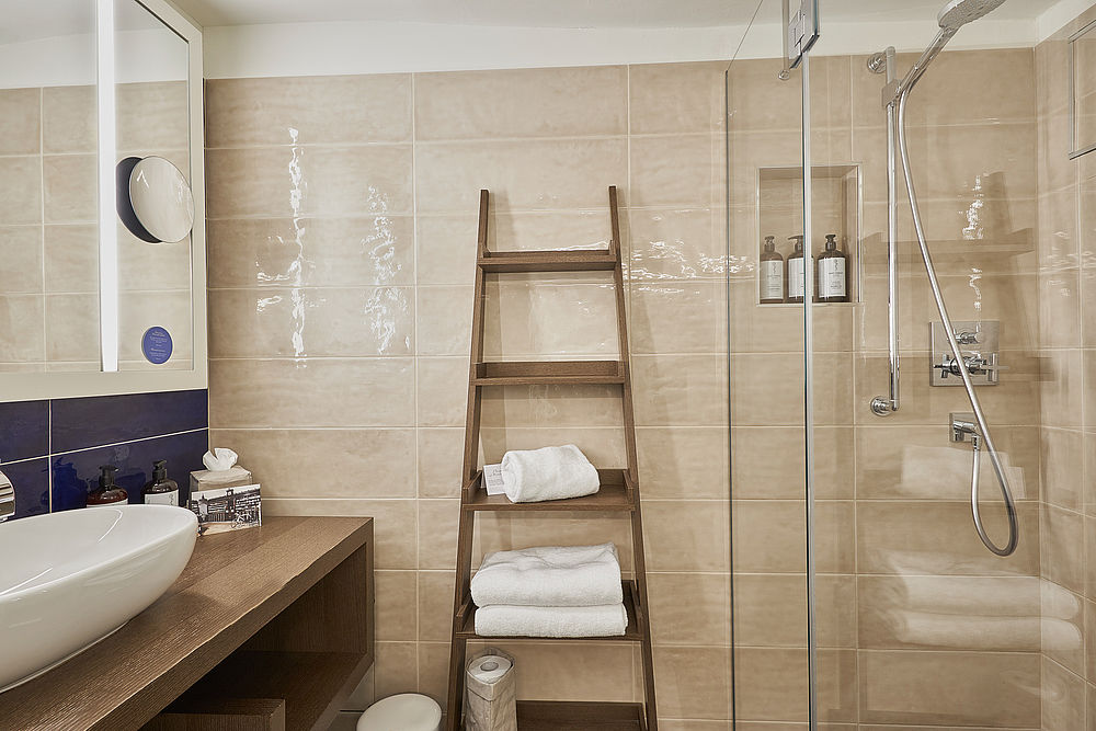 Badezimmer mit großer Dusche und Waschbecken, sowie champagner-farbenen Fließen und Holzleiter als Handtuchablage
