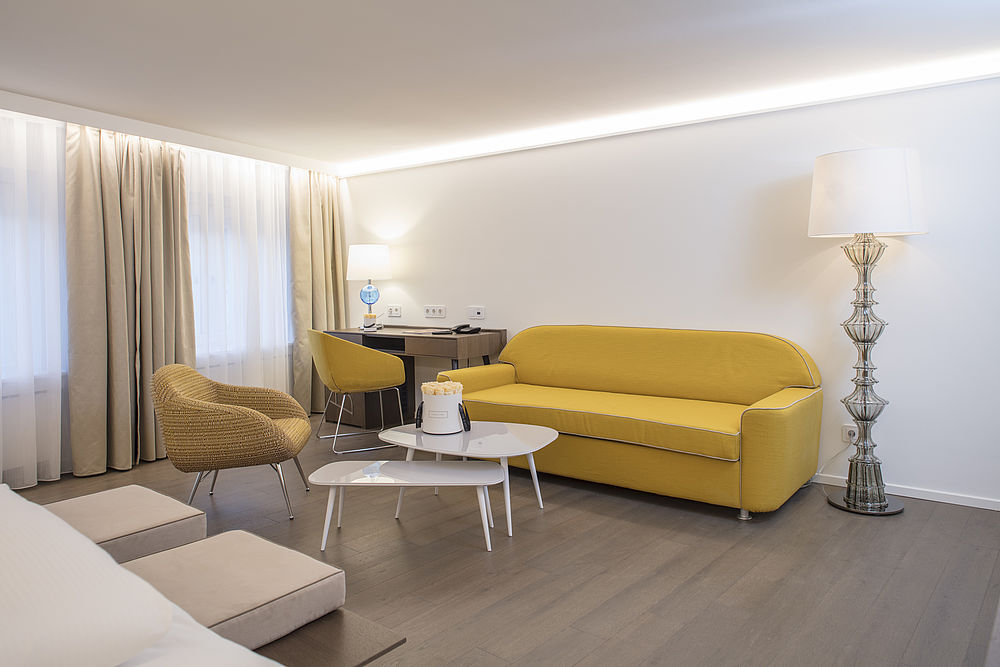 Der Wohnbereich einer Hotelsuite besitzt ein Sofa und Stühle aus Safran-Farben, beige Vorhänge eine verzierte Stehlampe und eine kleine Schreibtischecke