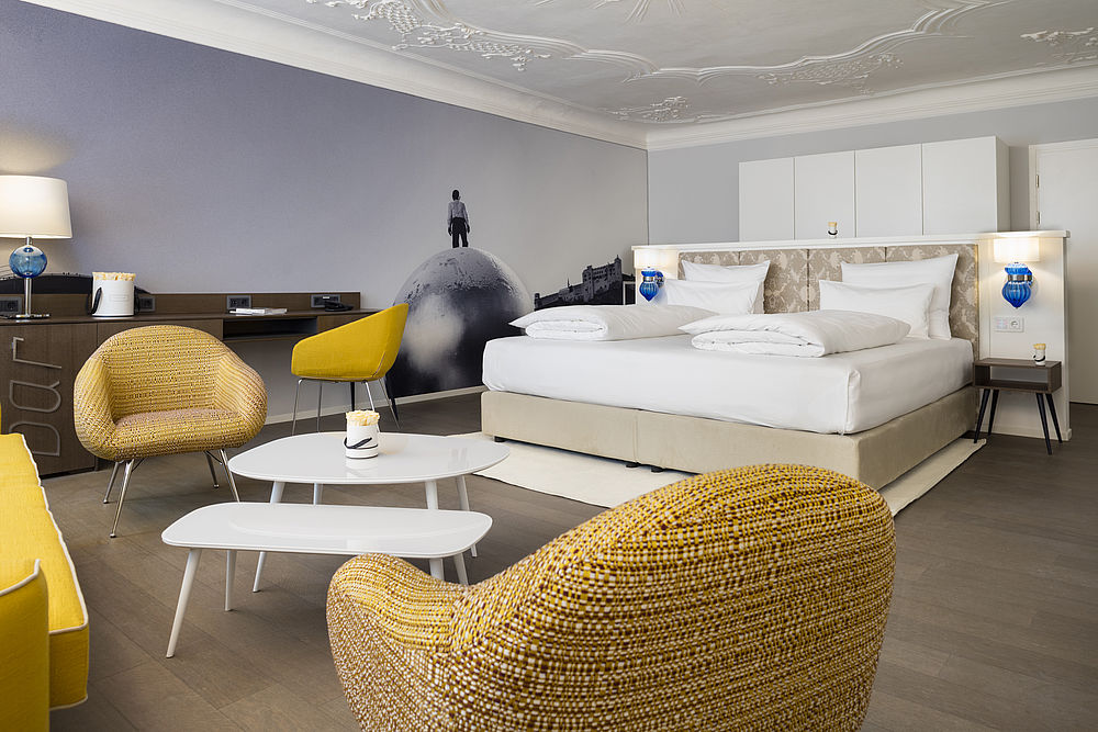 Das Schlafzimmer der Hotelsuite mit großem Boxspringbett, Sitzecke in Safran-Farben umgeben von möbeln im goldenen Design