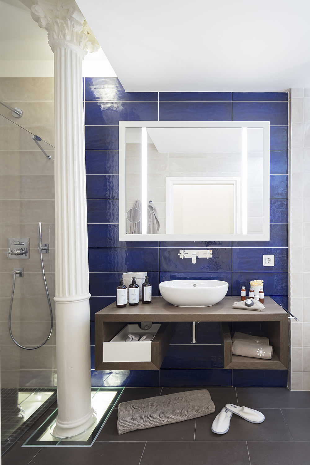 Das Badezimmer einer Suite mit blauen Fließen, weißer Säule und braunen Möbeln aus Holz