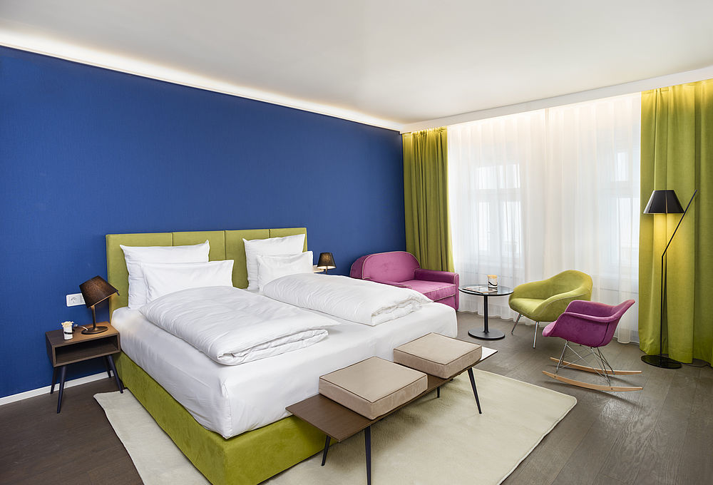 Junior Suite des Hotels mit grünem Bettrahmen und Vorhängen, königsblauer Wandfarbe und pinken Samtstühlen