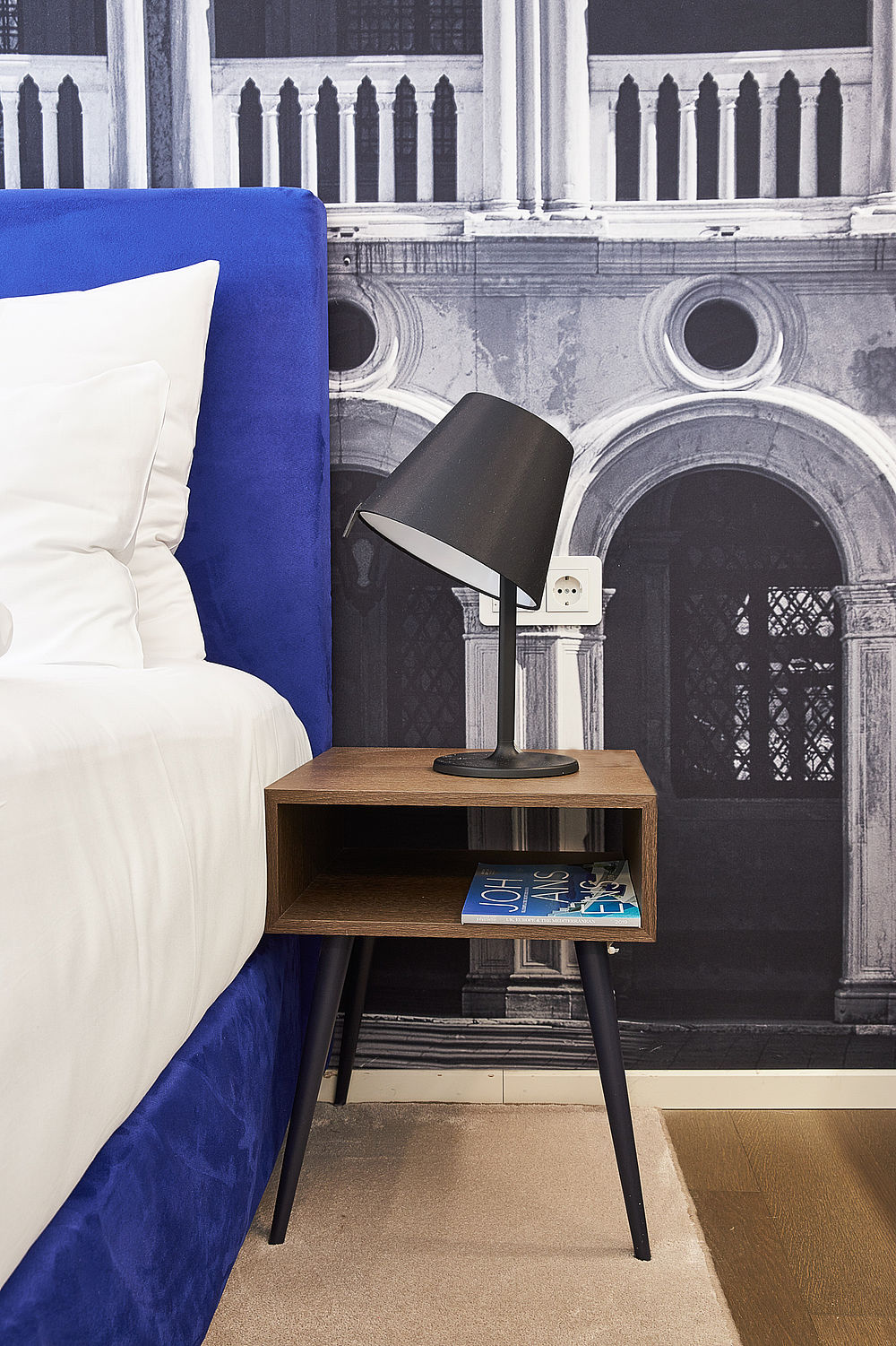 Details eines Hotelzimmers mit braunem Holznachttisch und schwarzer Lampe neben dem königsblauen Boxspringbett