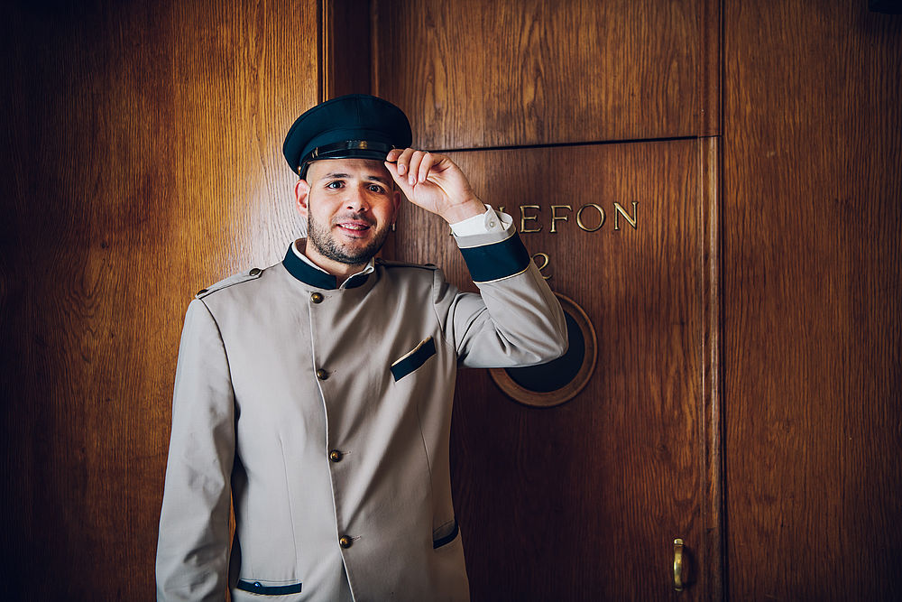 Ein Hotelmitarbeiter steht vor einer braunen Holztür in Uniform und Kapitänsmütze