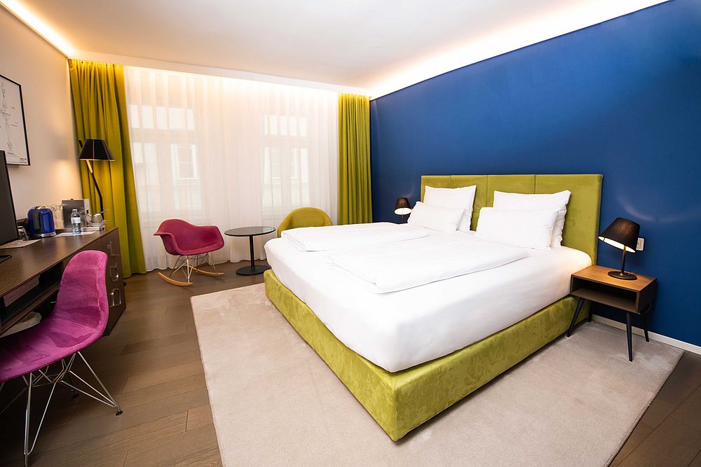 Gelbes Boxspringbrett vor blauer Wand und Sitzecke mit Schaukelstühlen in kräftigen Farbentönen in der Junior Suite des Hotel Stein
