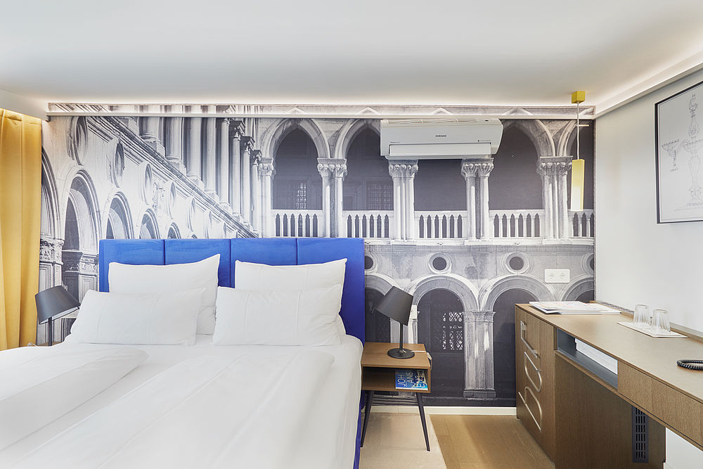 Ein Hotelzimmer mit großem Doppelbett, Ablagefläche und schwarz-weiß Fotografie als Wandtapete