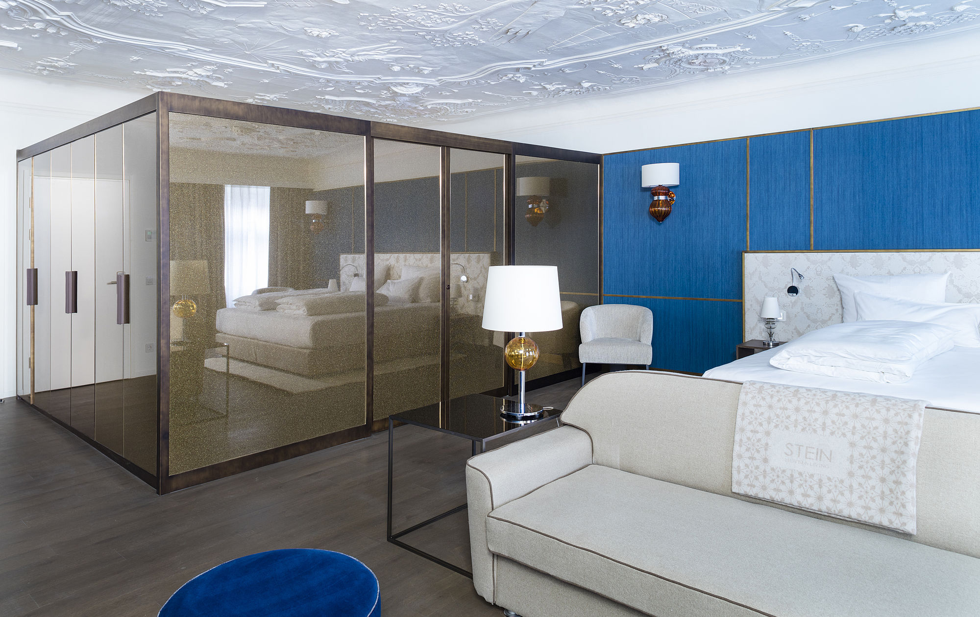 Honeymoon-Suite des Hotel Stein mit Stuckdecke, Kingsize-Bett vor blauer Wand mit goldenen Aktzenten, beigem Sofa und geräumigem Metallschrank mit Spiegelwand