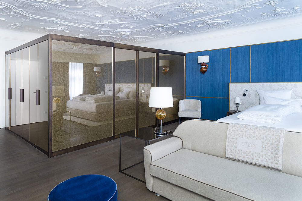 Suite luna di miele dell'Hotel Stein con soffitto a stucco, letto king-size davanti a una parete blu, divano beige e ampio armadio in metallo