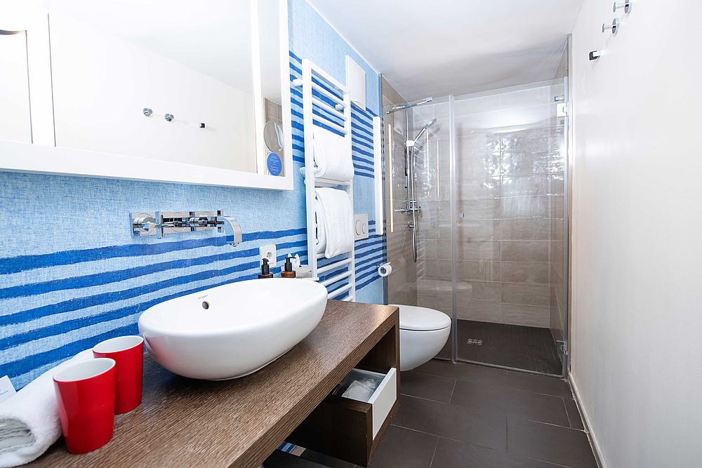 Bad der Junior Suite im Hotel Stein mit hellblauer Wand, Waschtischkonsole aus dunklem Holz,  Naturkosmetik und weißen Handtüchern und einer Glas-Duschkabine