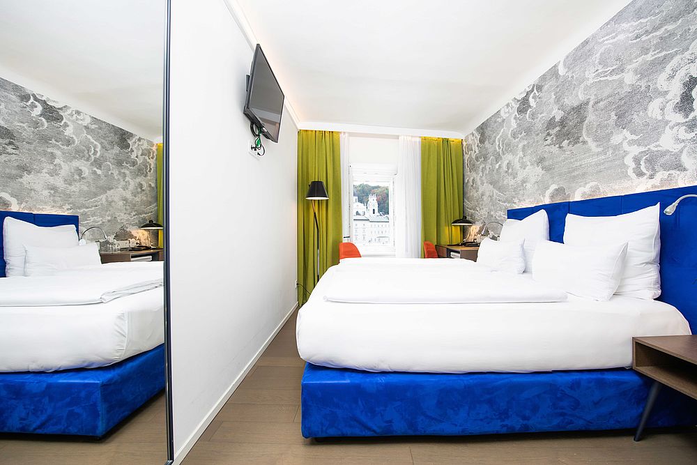 Doppelzimmer des Designhotel Stein mit blauem Boxspringbett, Flachbildschirm, Schreibtisch am Fenster und großter Spiegelwand