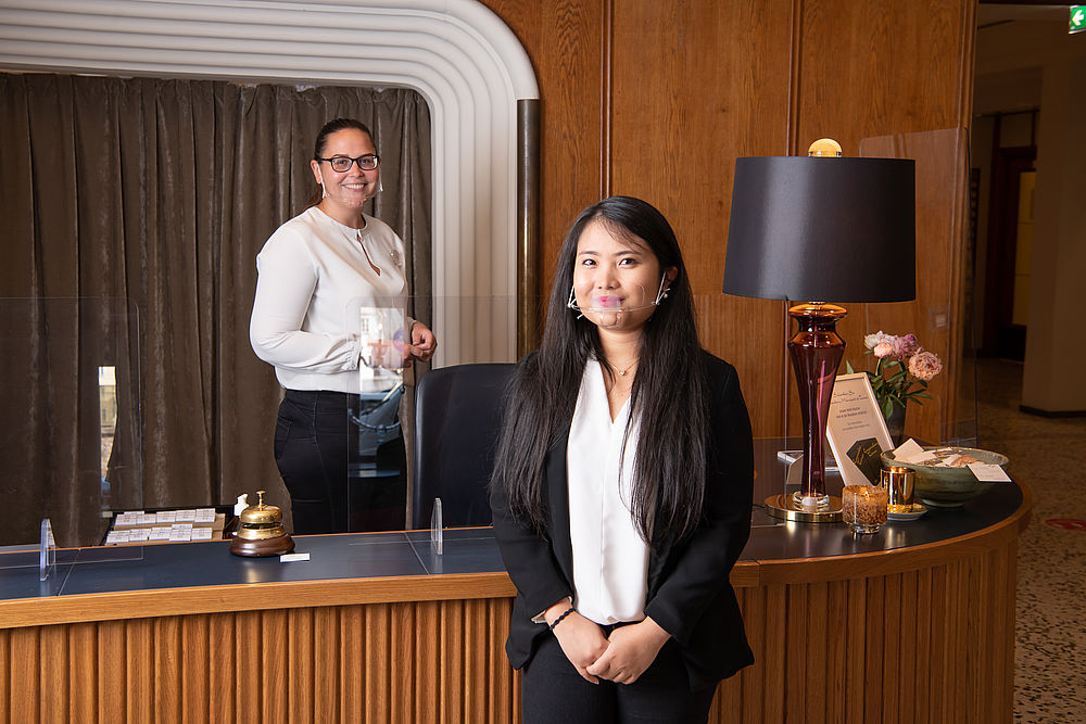 Zwei Rezeptionistinnen stehen lächelnd am Empfang des Hotels mit Mund-Nasen-Bedeckung